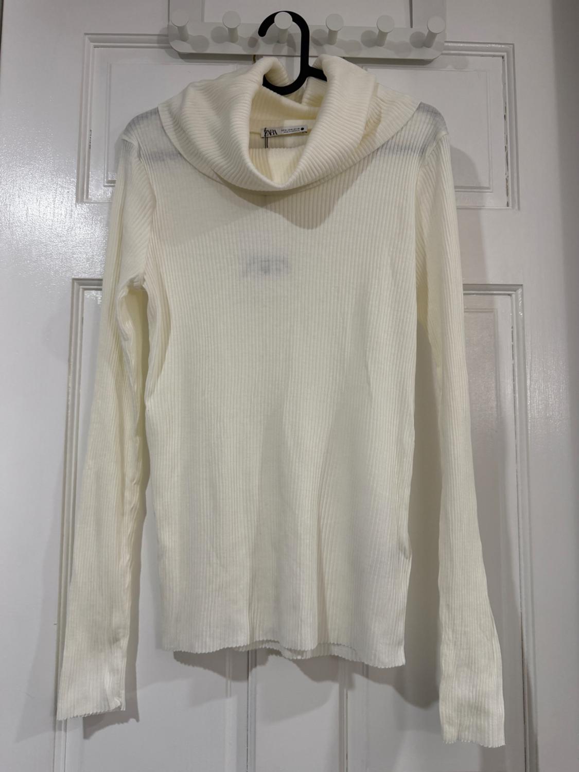 White ribbed knit roll-neck jumper, hanging on a black hanger over a white door.