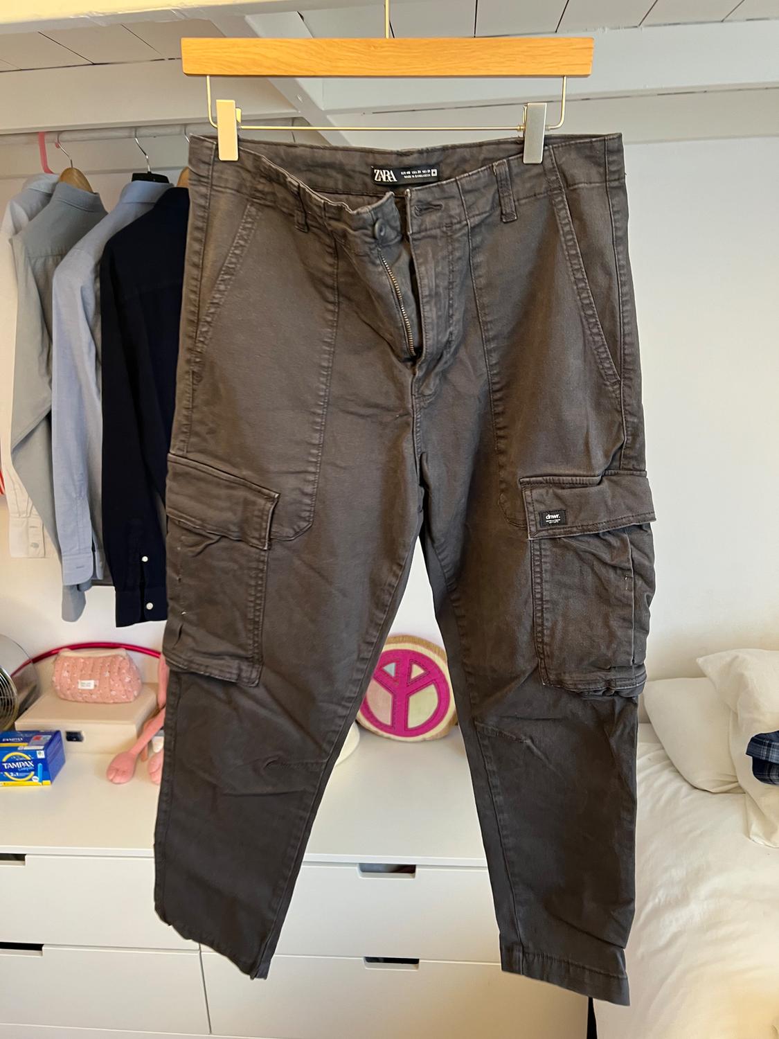 Grey cargo trousers hanging on a hanger, with side pockets. Background with clothes hanging and objects on a white chest of drawers.