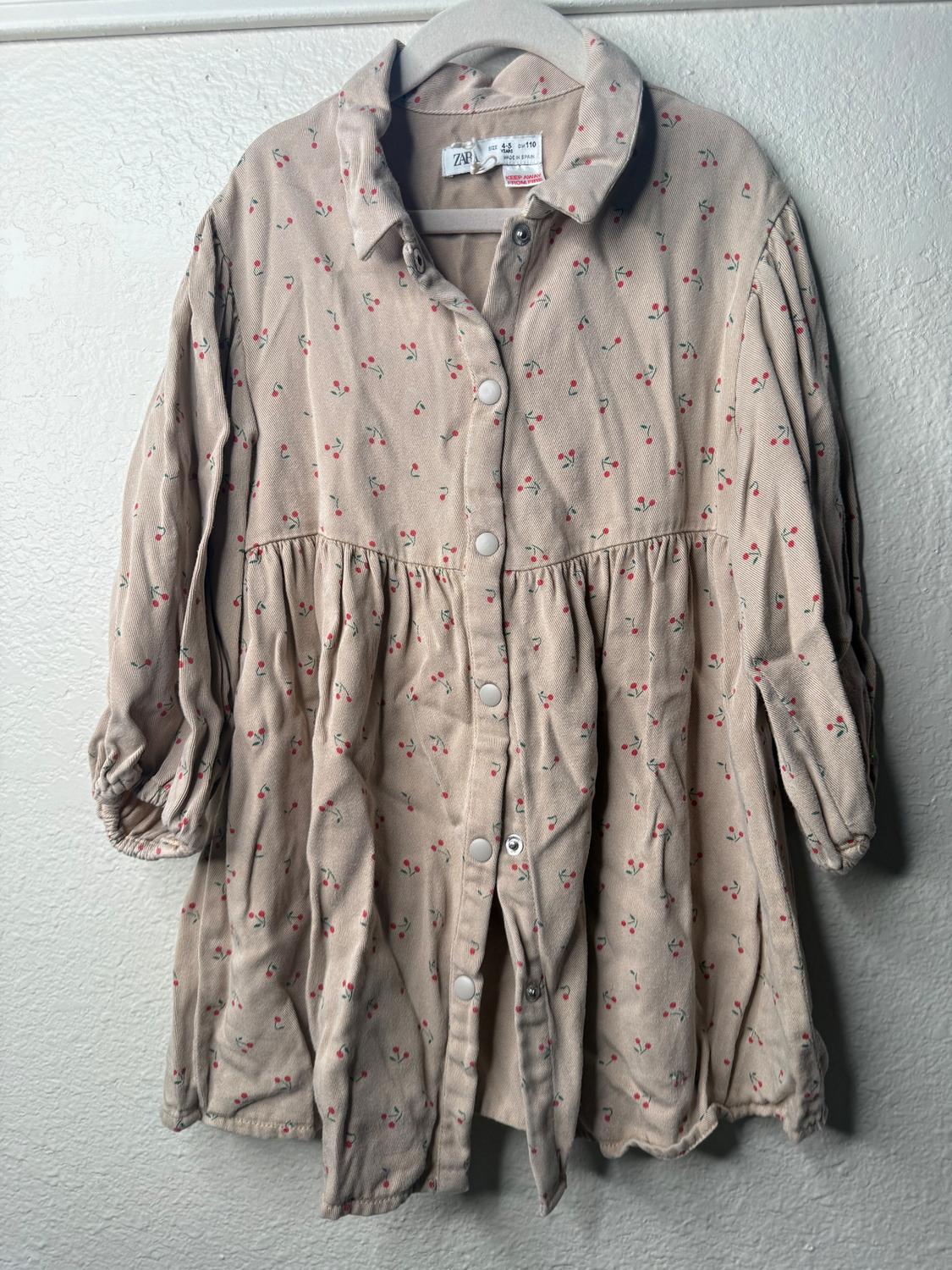Beige shirt with a small red and green floral print, hanging on a white hanger against a light-colored wall.