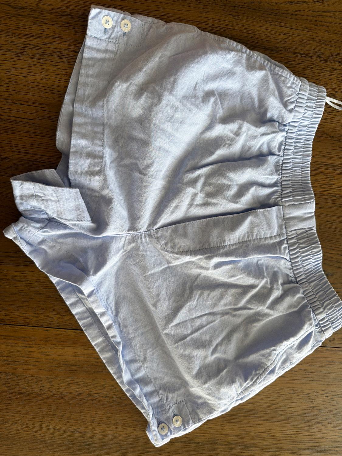 Light blue casual shorts with an elasticated waistband and buttons at the hem, laid out on a wooden surface for a relaxed look.