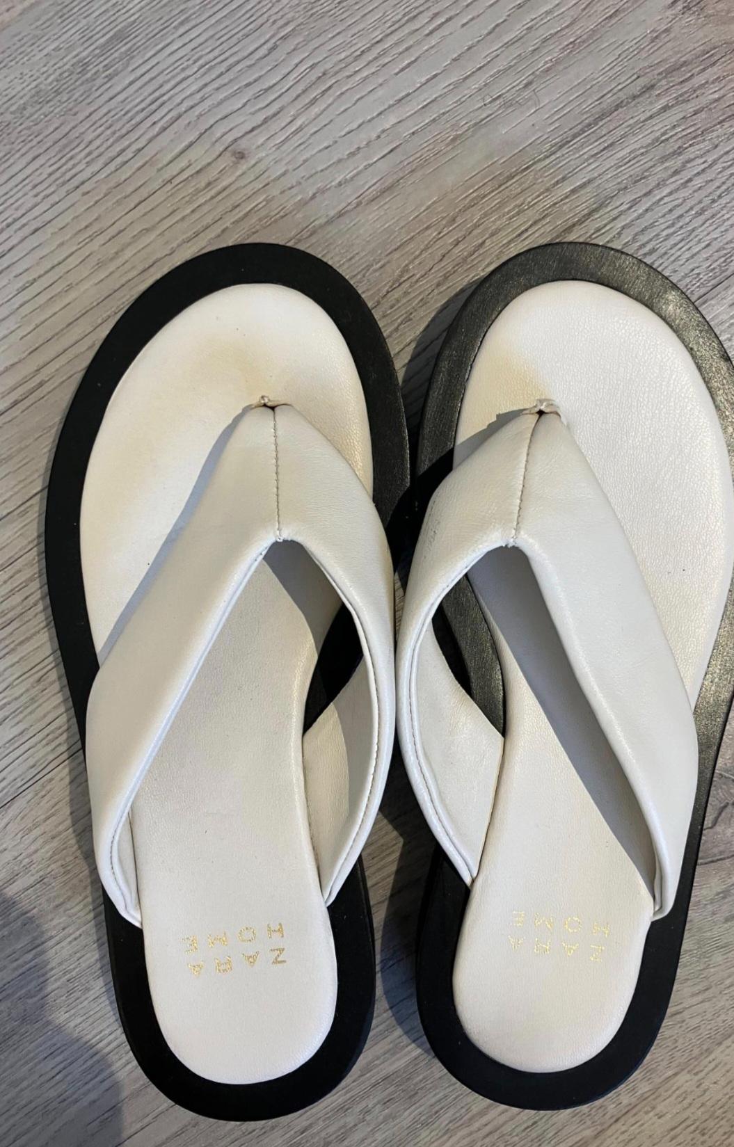 Pair of white strappy sandals with a white insole on a thick black sole on a wooden floor. Gold ZARA HOME text on the insole.