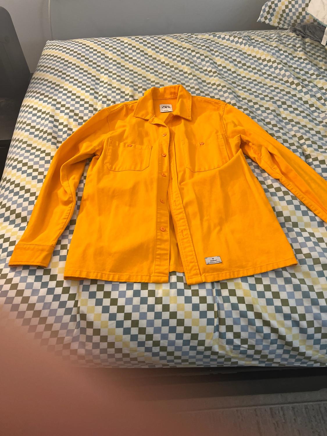 Yellow long-sleeve button-down shirt with pockets, on a bed with plaid sheets. Tag with ZARA text on the collar.