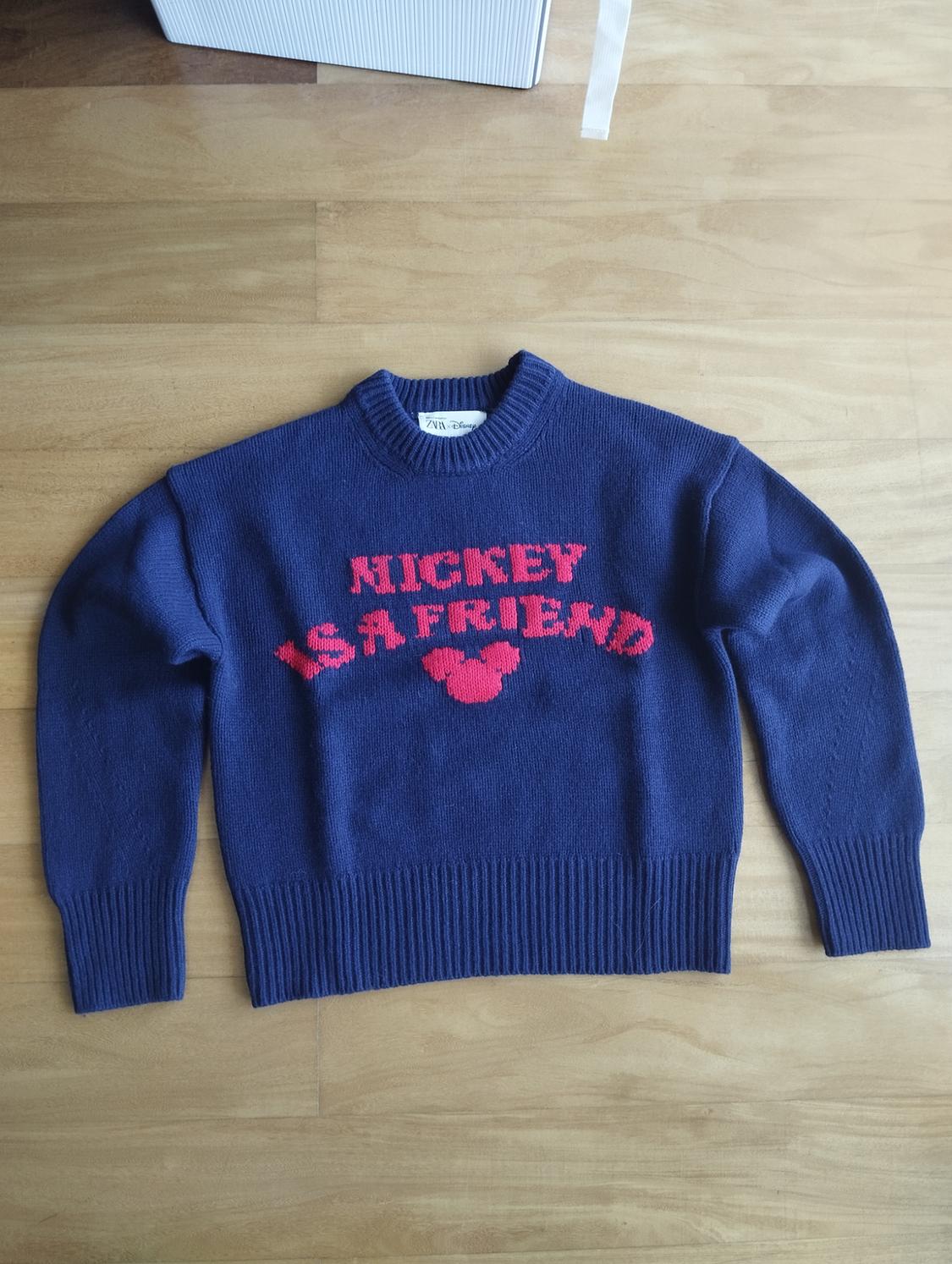 Blue jumper with red text "MICKEY IS A FRIEND" and a Mickey Mouse icon in the centre, on a light wooden floor.