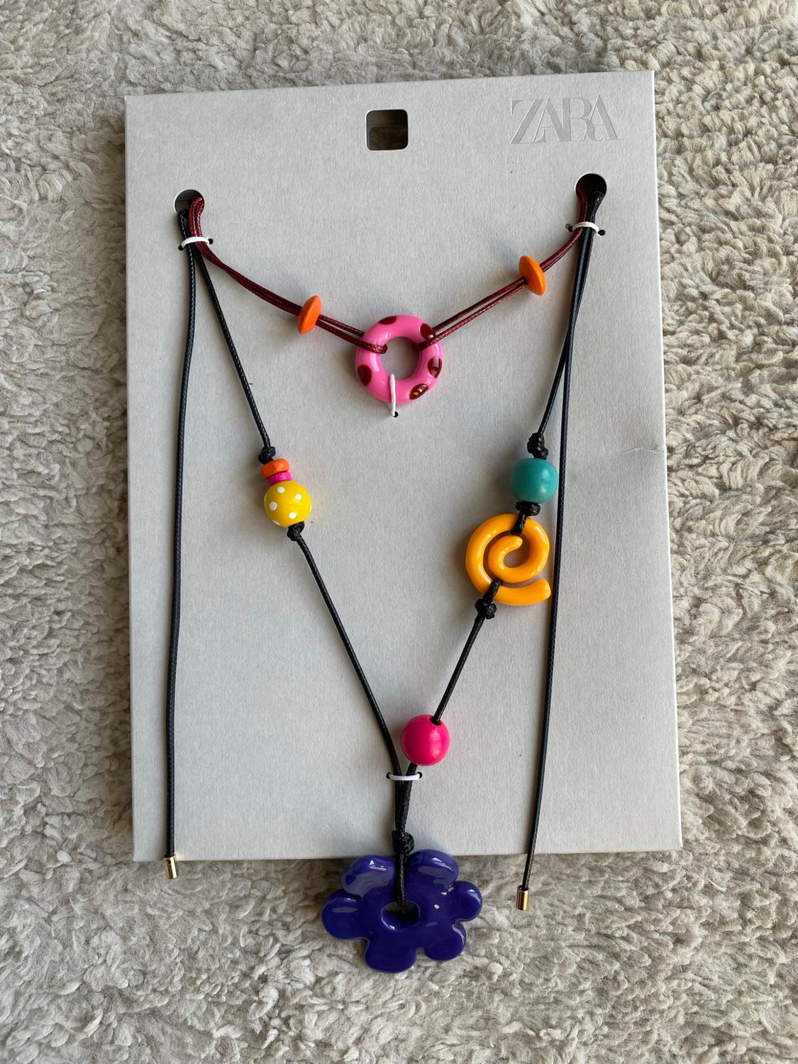 Two necklaces with colourful beads, a pink hoop and a purple flower on card bearing the text ZARA, against a softly textured background.