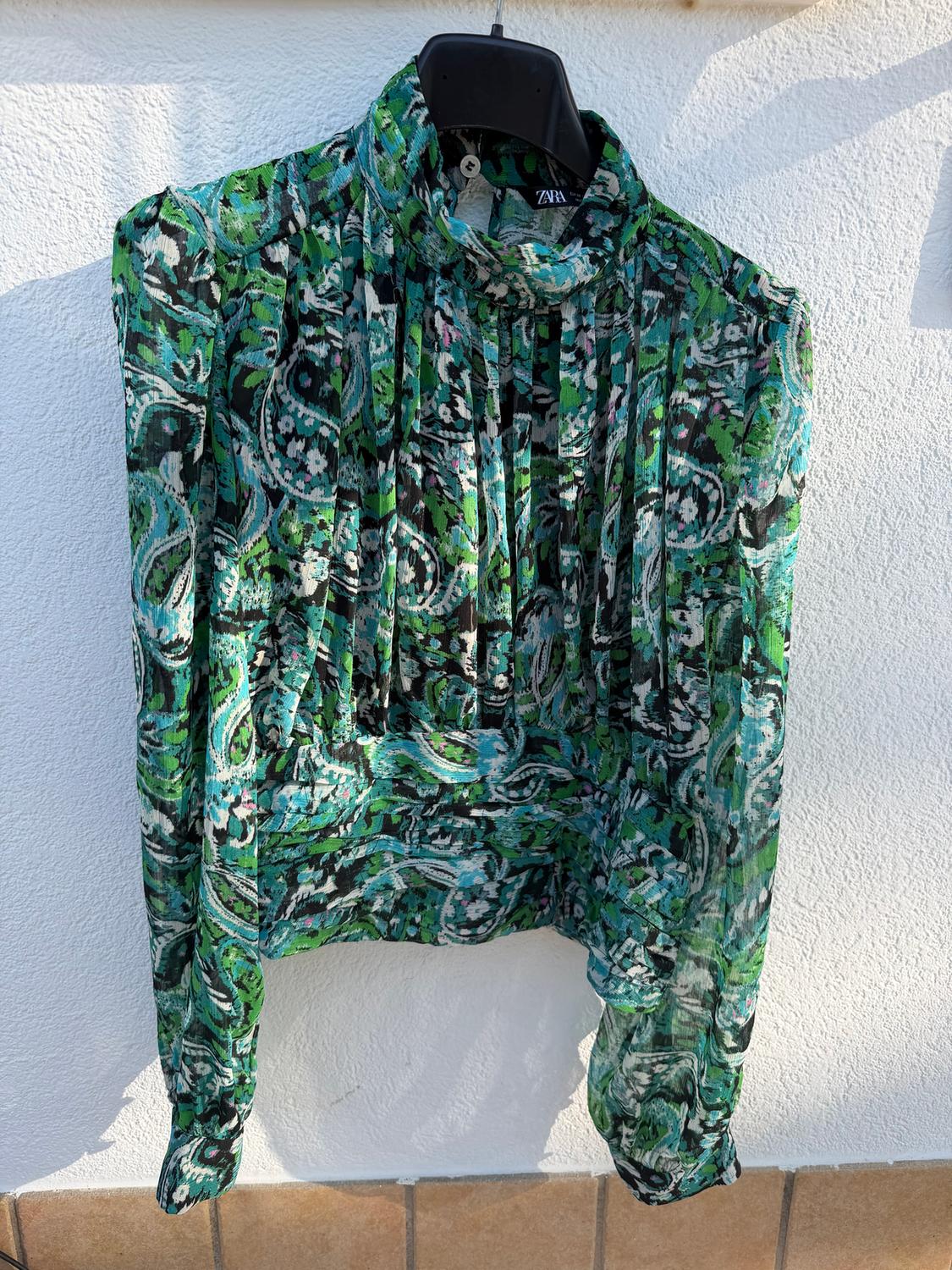 Patterned blouse in shades of green and black, hanging on a black hanger against a sunlit white wall.