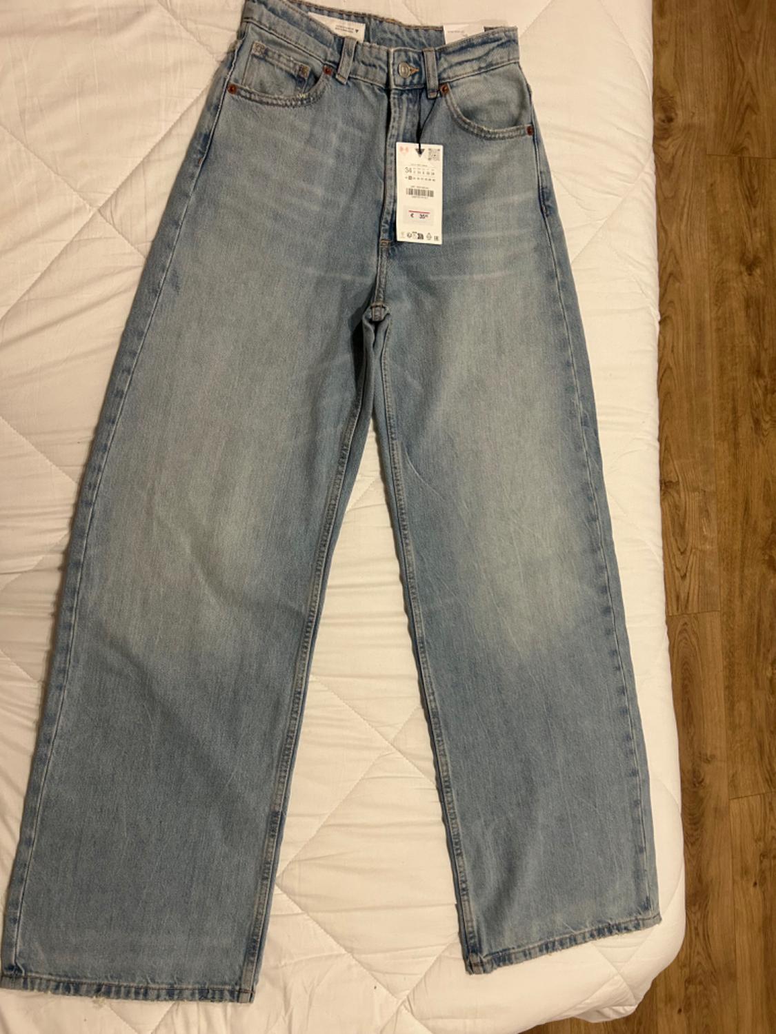 Light blue denim jeans with a visible label, placed on a white bedspread and wooden floor.