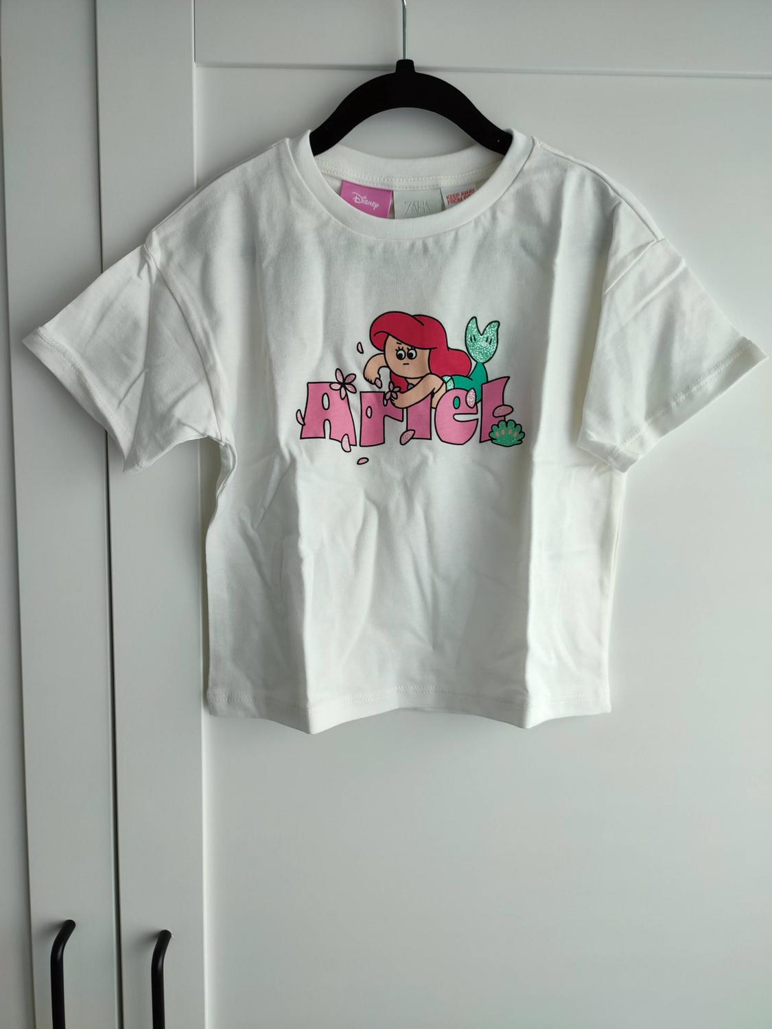 White T-shirt on a black hanger above a white wardrobe. Mermaid print and Ariel text, Disney, zara, KEEP AWAY FROM FIRE.