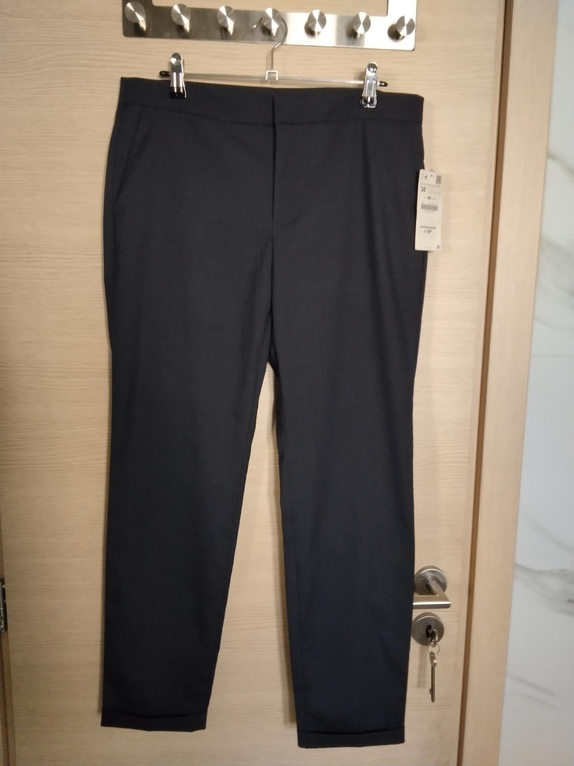 Dark trousers hanging from a hanger over a light wooden door. The garment has a label, and a metal door handle can be seen underneath.