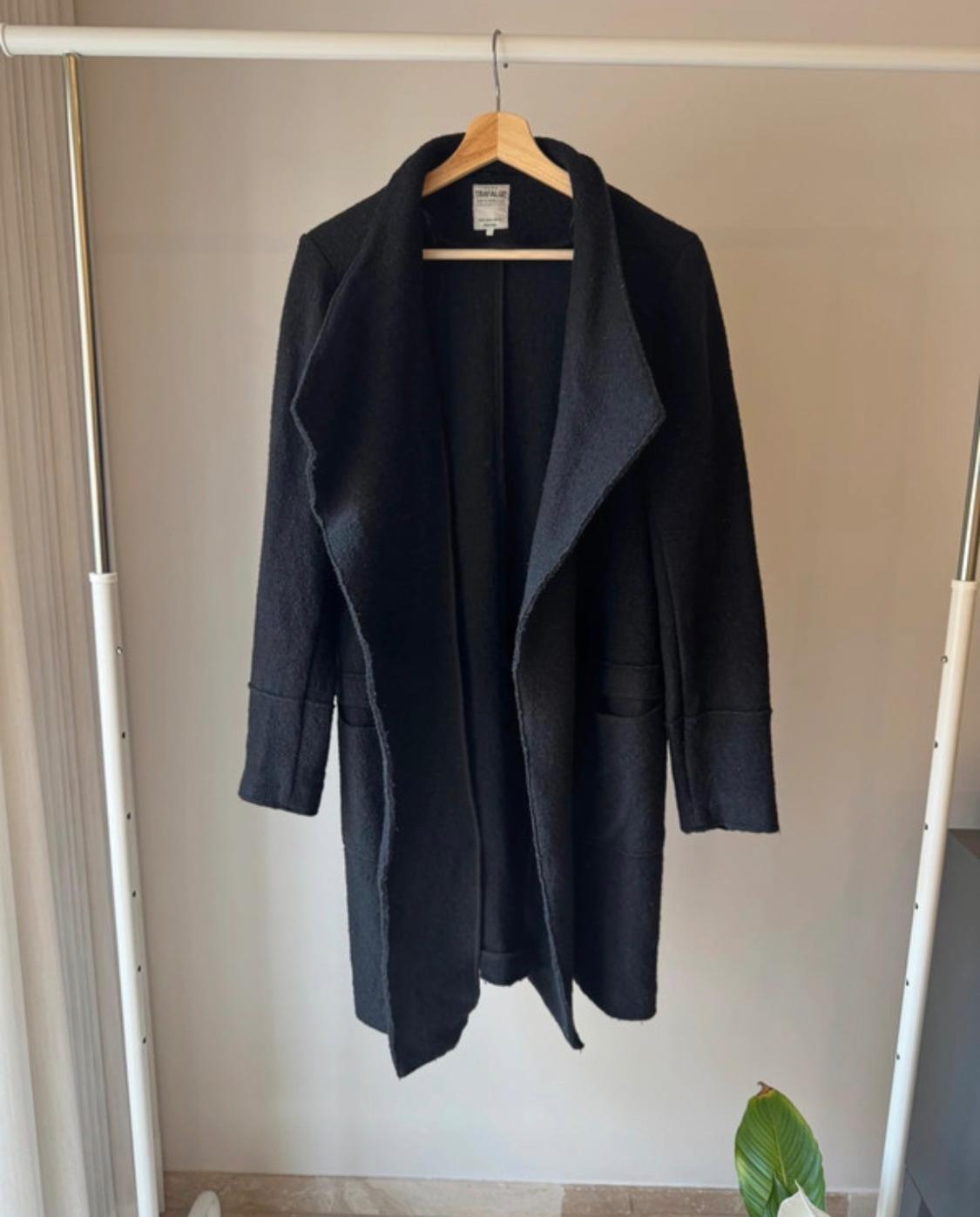 Black coat hanging on a white coat rack, with a beige wall background and a green plant in the bottom right corner.