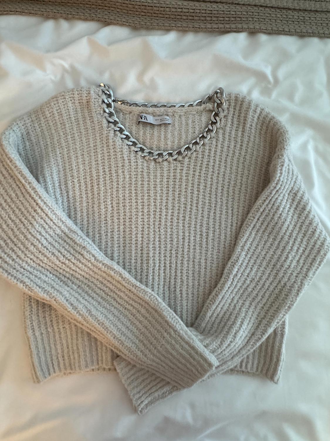Beige knitted jumper with a chunky silver chain on the collar, on a white bed with a brown bedspread. Visible label: "Zara".