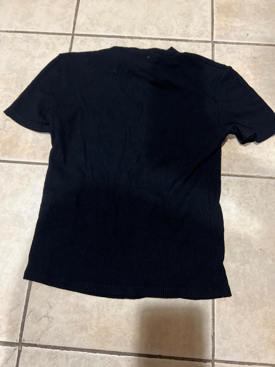 Black short-sleeved T-shirt on a beige tiled floor. The fabric appears ribbed and is laid out face down.