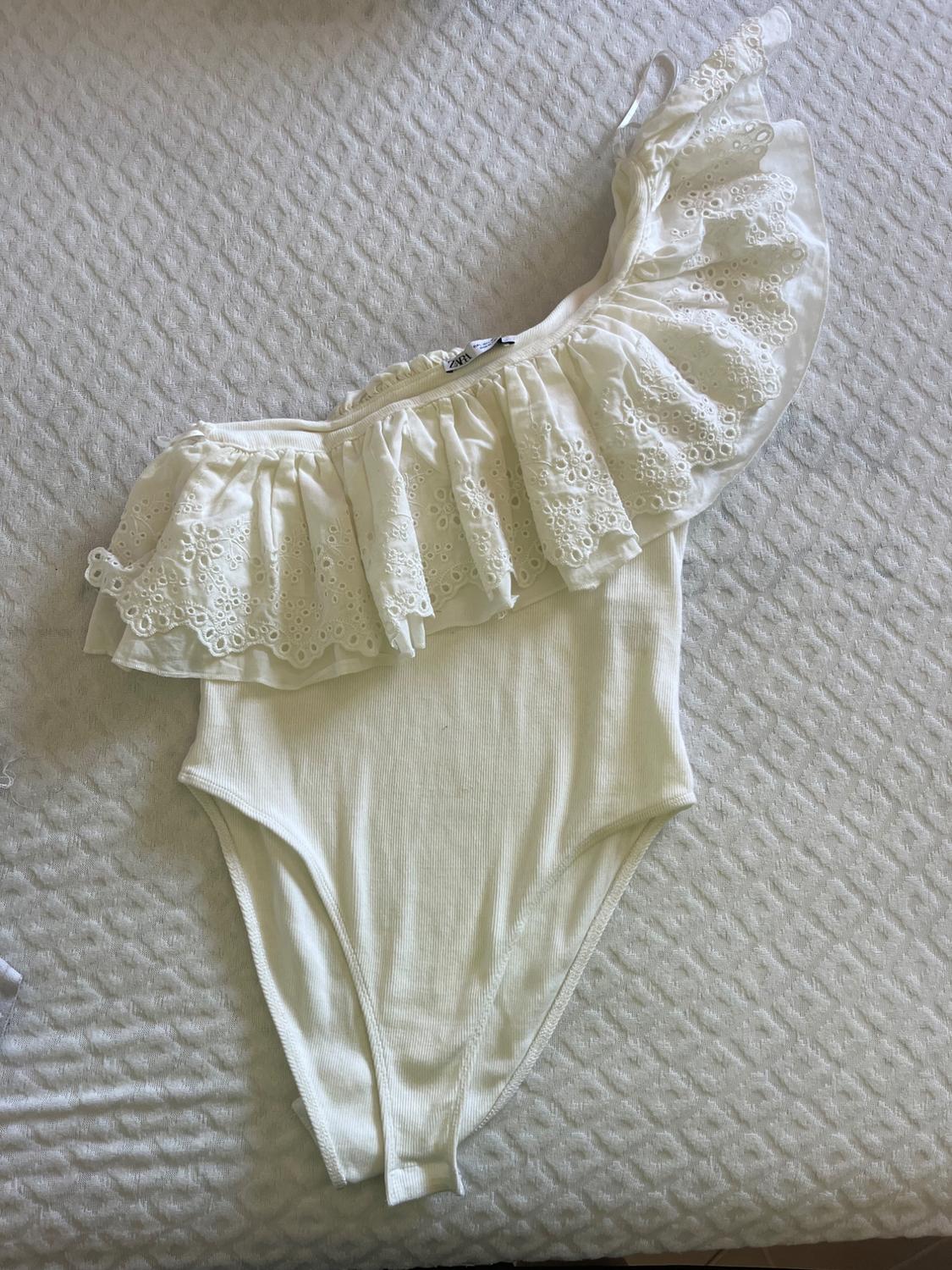 Ribbed white bodysuit with an asymmetric neckline and a ruffled trim, on a textured surface. Label: ZARA.