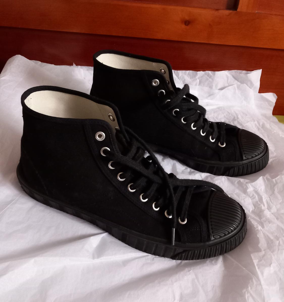 Black canvas lace-up trainers with rubber soles, on white paper. Brown wooden background.