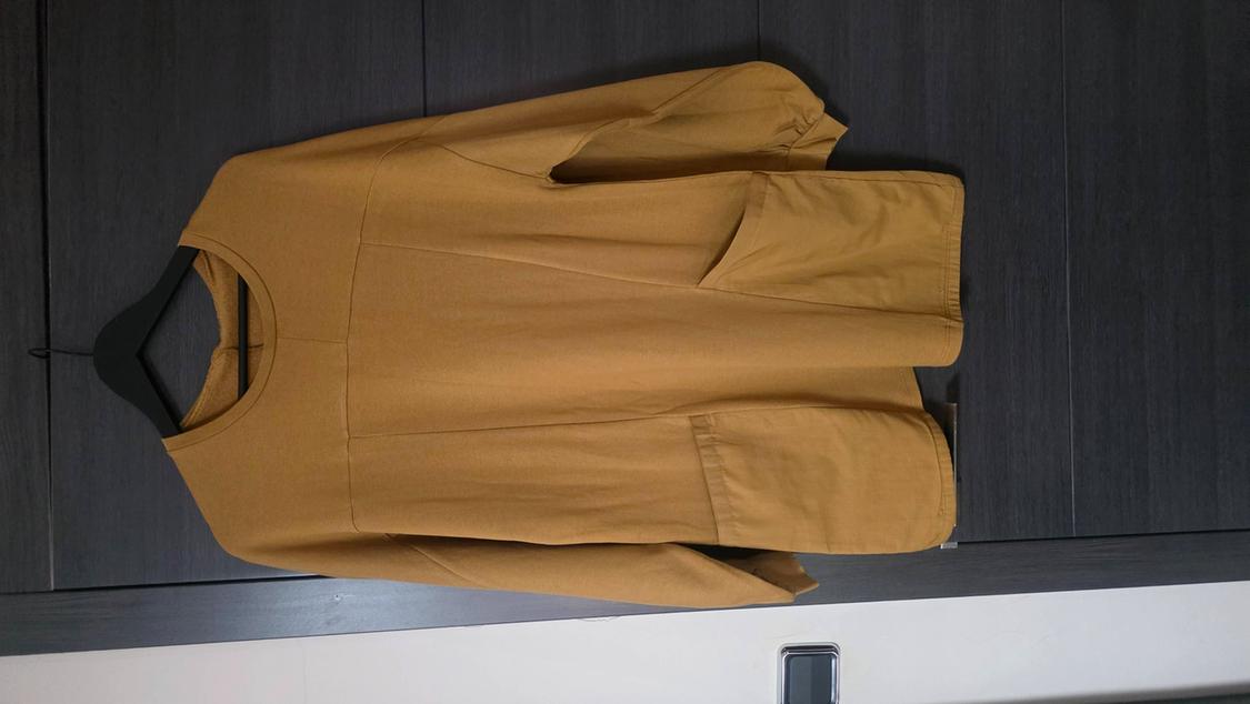 Mustard-coloured long-sleeved, round-neck overshirt with large pockets, hanging on a black hanger against a dark wood background.