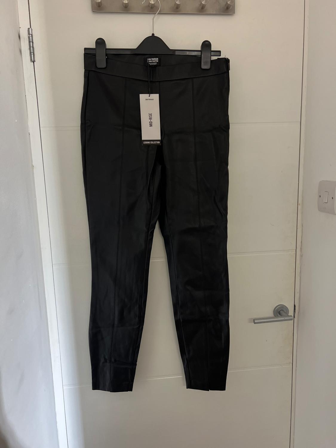 Black trousers hanging on a hanger over a white door. Visible tag with text: "MID-RISE LEGGING COLLECTION".