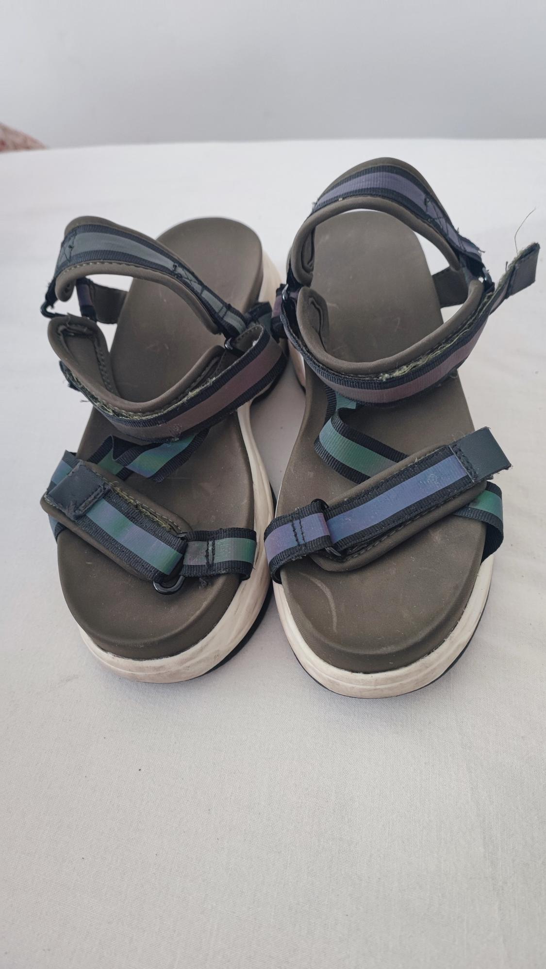 Black strappy sandals with white soles on a white surface. The straps have blue and green detailing.