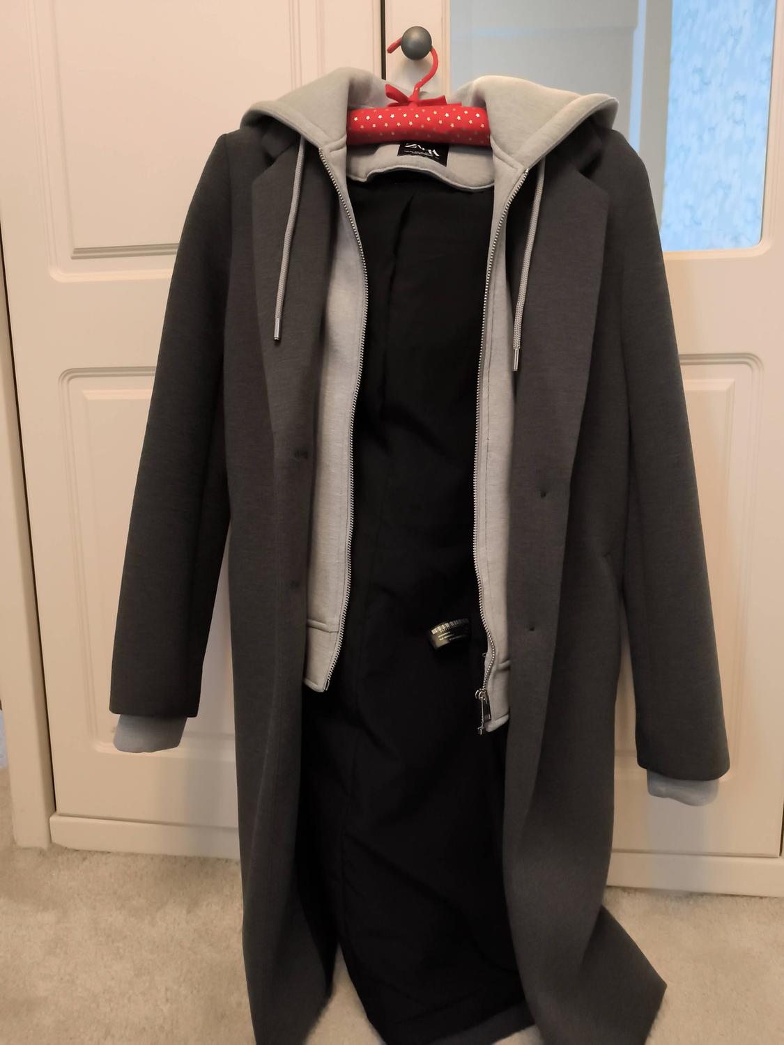 Grey coat with a light grey hood and zip, hanging on a red hanger with white dots, in front of a white door.