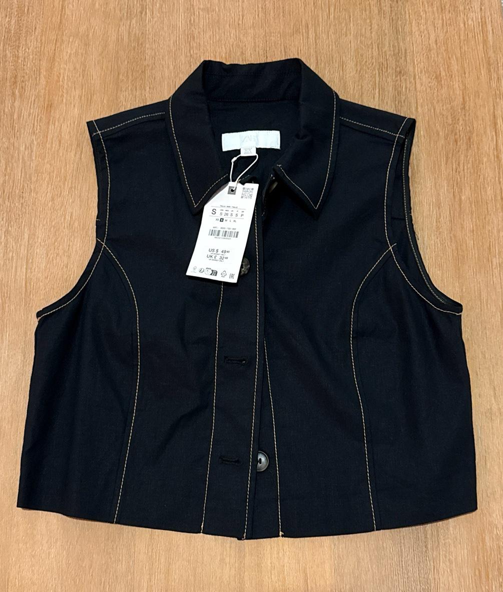Black vest with collar, buttons, and contrast stitching on wood. Label with text ZARA S US S 49.90.