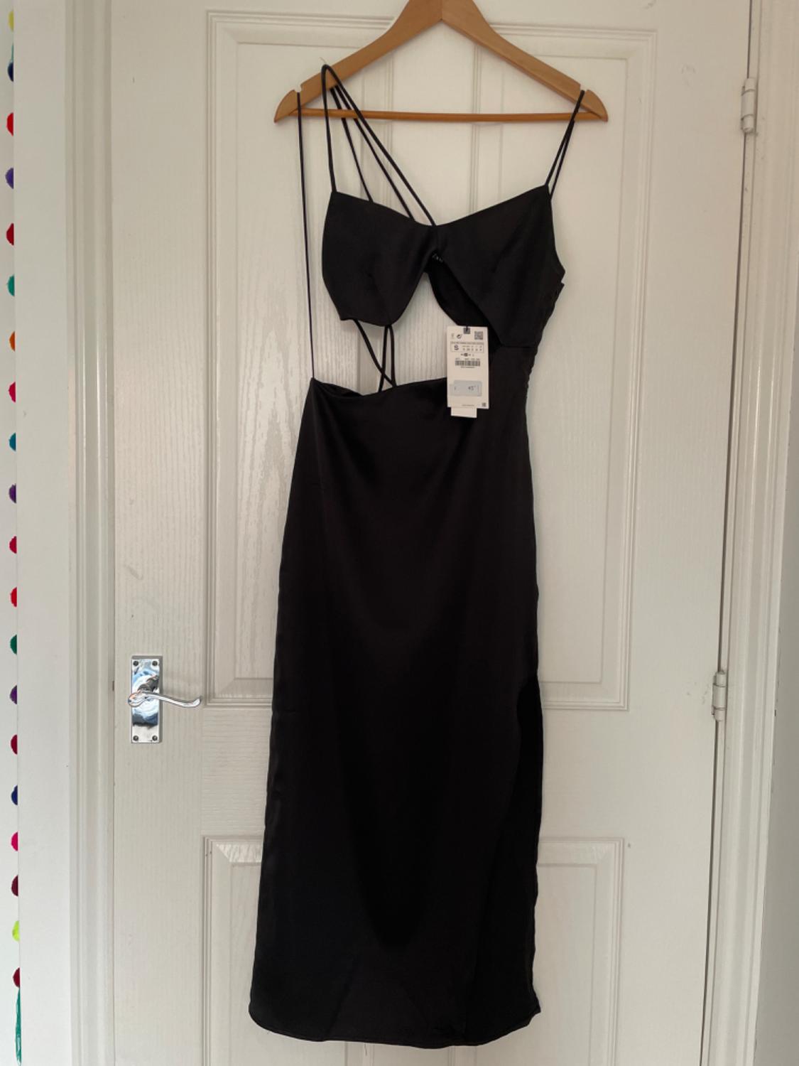 Black dress with cut-outs, asymmetrical spaghetti straps and label, hanging on a wooden hanger on a white door in a casual setting.