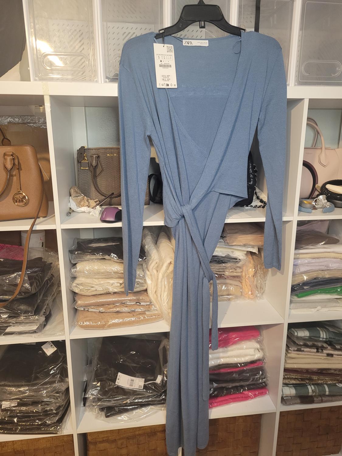 Long-sleeved blue dress hanging on a rack in front of shelves with handbags and clothes folded in plastic bags.