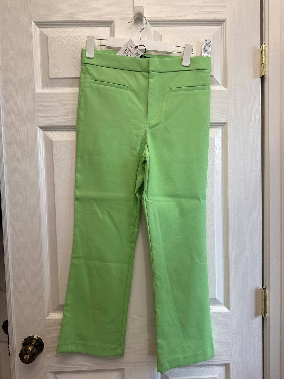 Green trousers hanging on a white door with a hanger. Visible front pockets and swing tag.