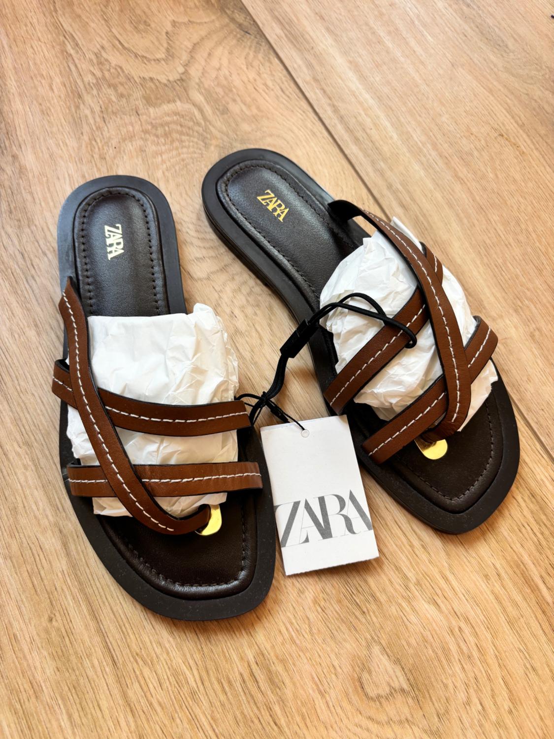 Brown flat sandals with crossover straps and white stitching on wood, with stuffing paper. Text on insole and label: ZARA.