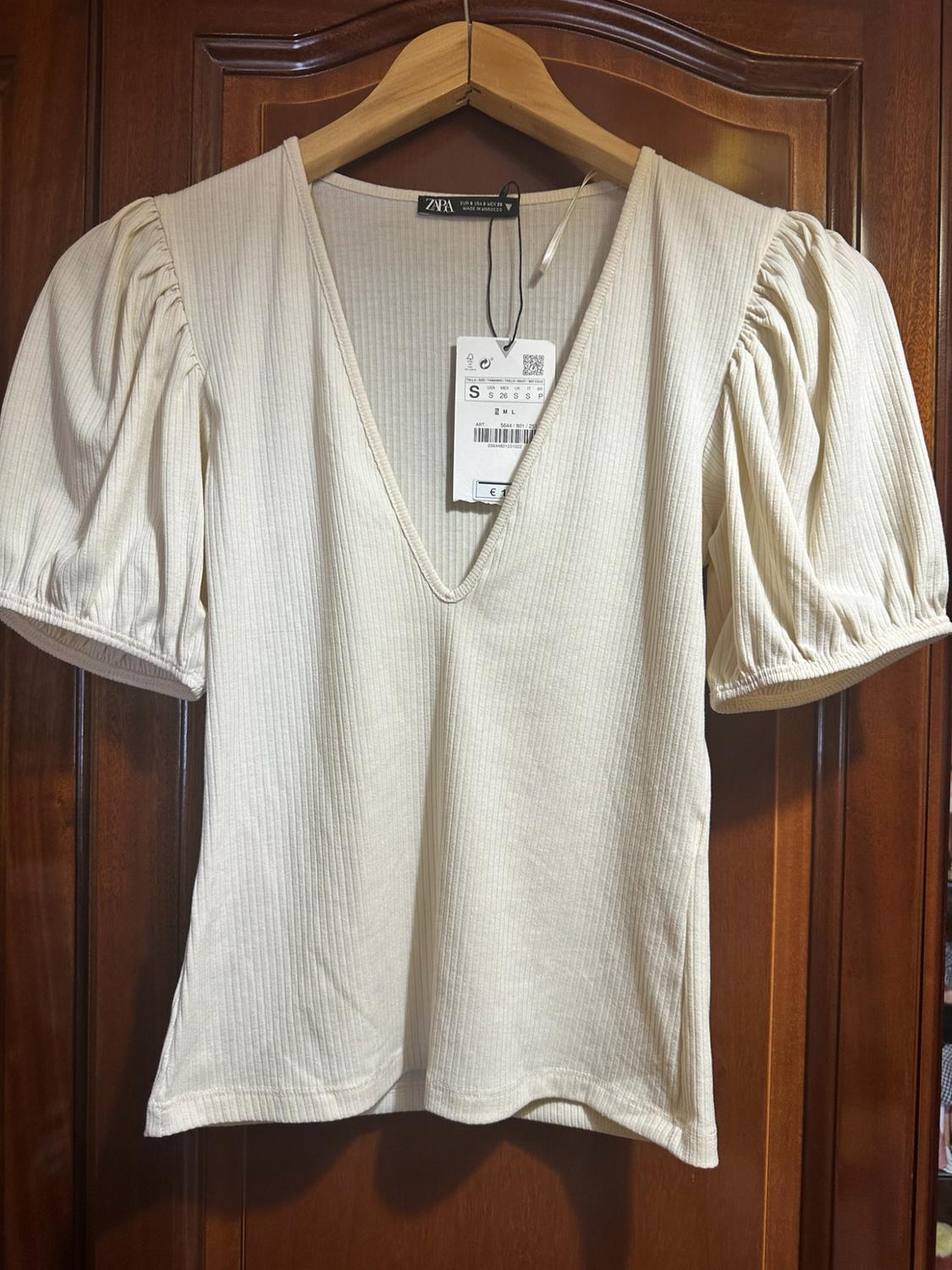 Ribbed cream top with V-neck and puffed sleeves, hanging on a wooden hanger. Labels with text: ZARA, S.