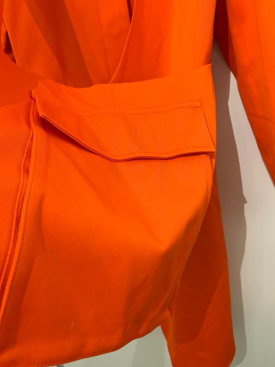 Detail of an orange jacket with a large sleeve pocket, highlighting the fabric and stitching on a neutral background.