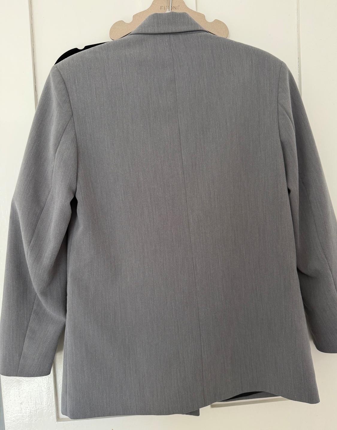 Back view of a grey blazer hanging on a wooden hanger with the text FIONI, against a white door. Minimalist style.
