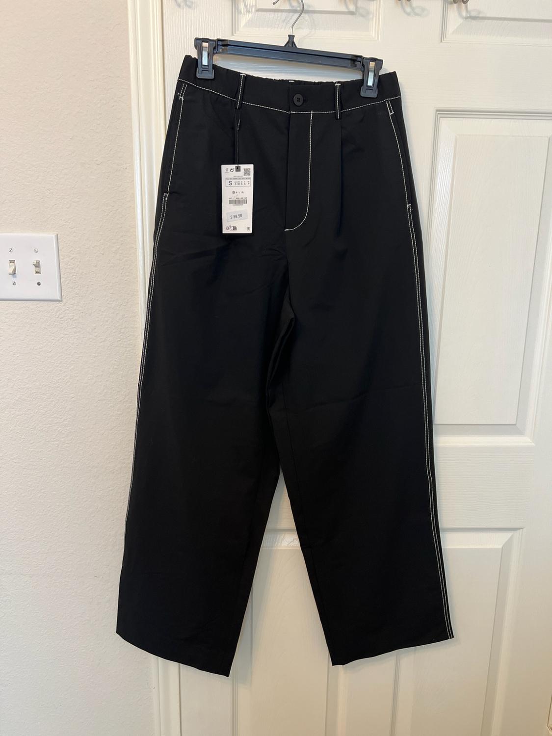 Black trousers with white stitching hanging from a coat hanger on a white door. Visible text: S $69.90.