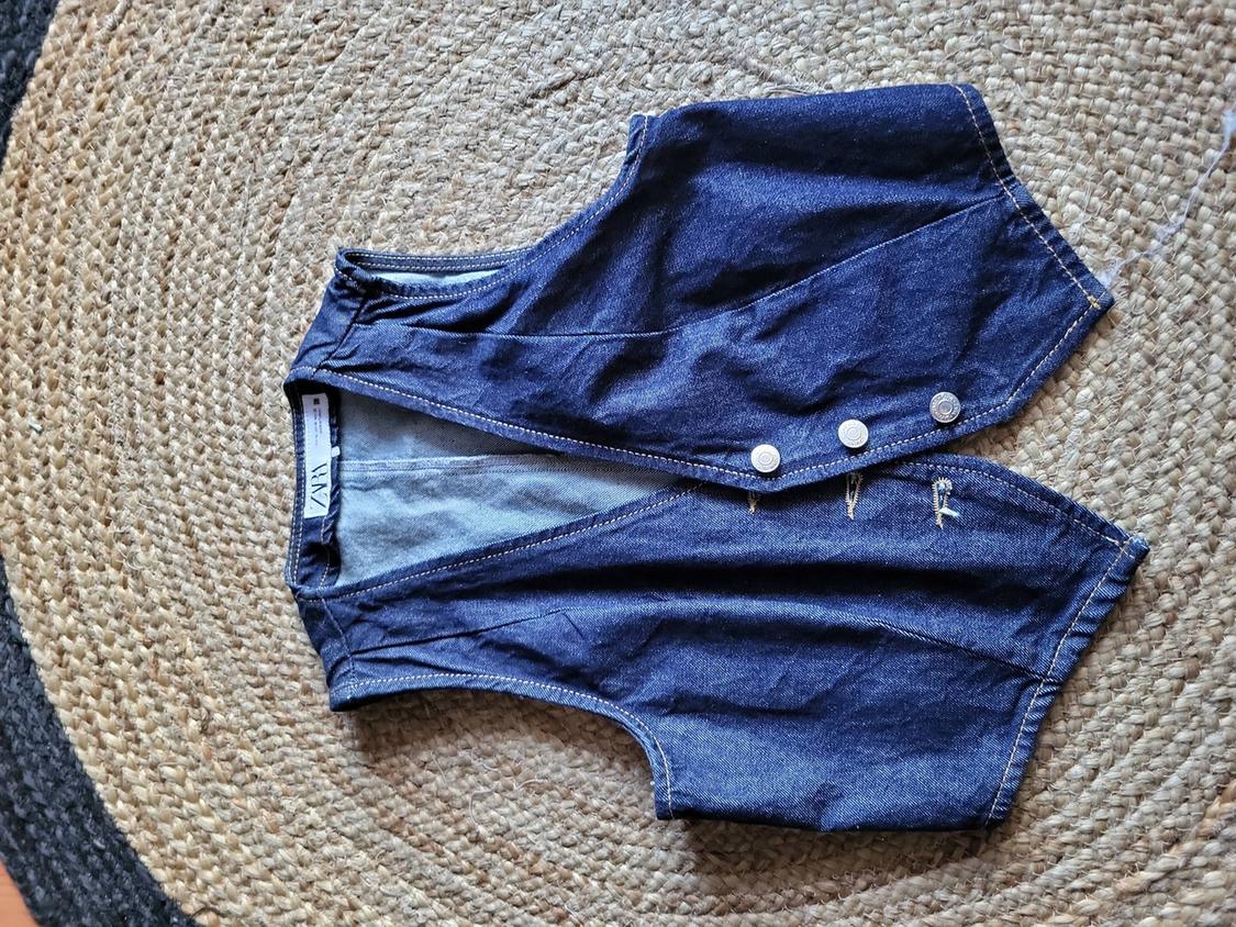 Blue denim waistcoat with silver buttons on a circular woven rug. Visible label with "ZARA" text.