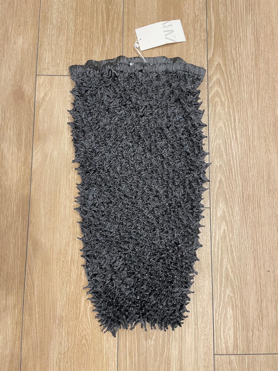 Black skirt with crinkled texture on light wooden floor. White label with 'ZARA' text attached at the top.