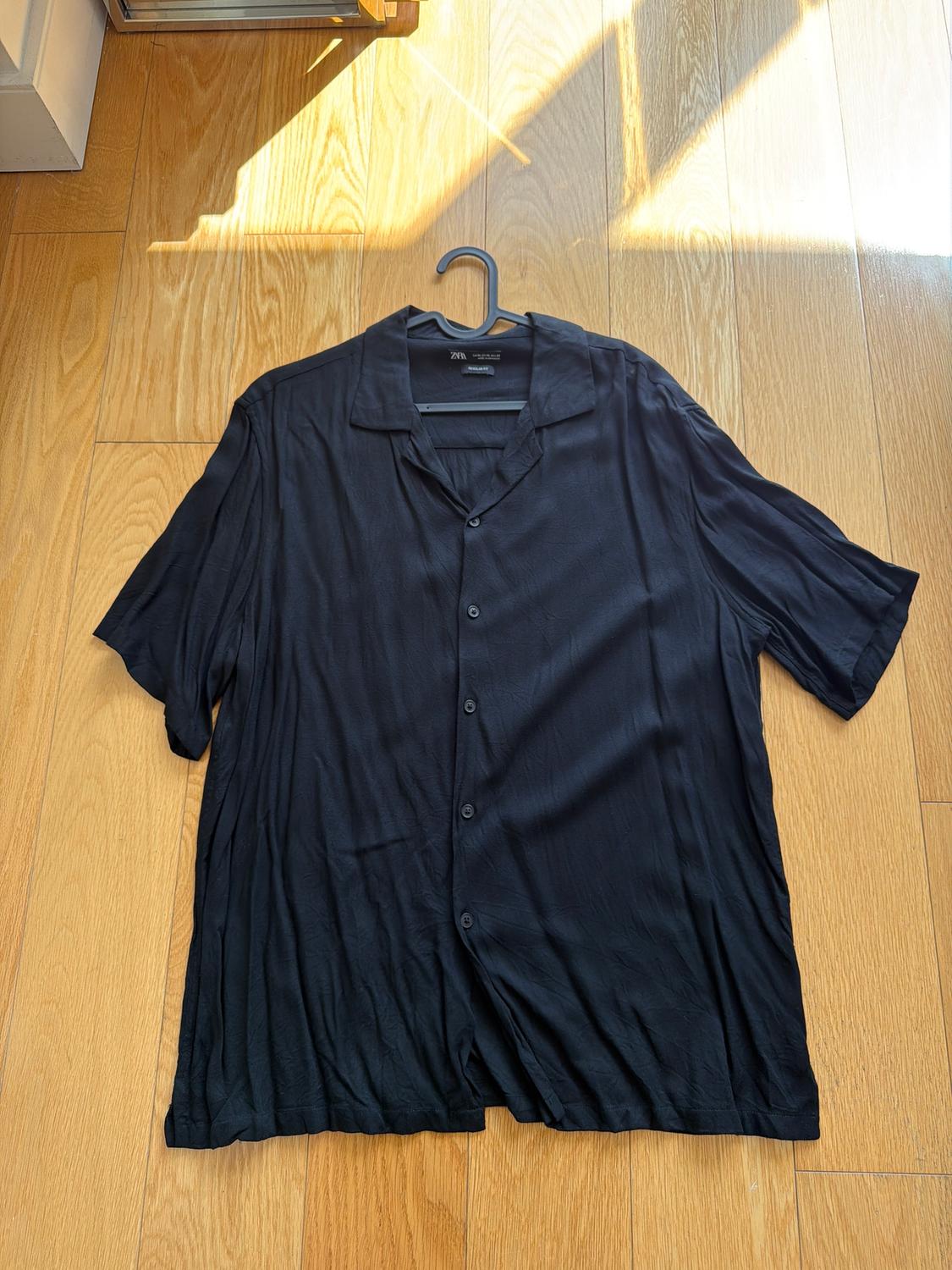 Black short-sleeved button-up shirt, hanging on a hanger over a light wood floor lit by sunlight.
