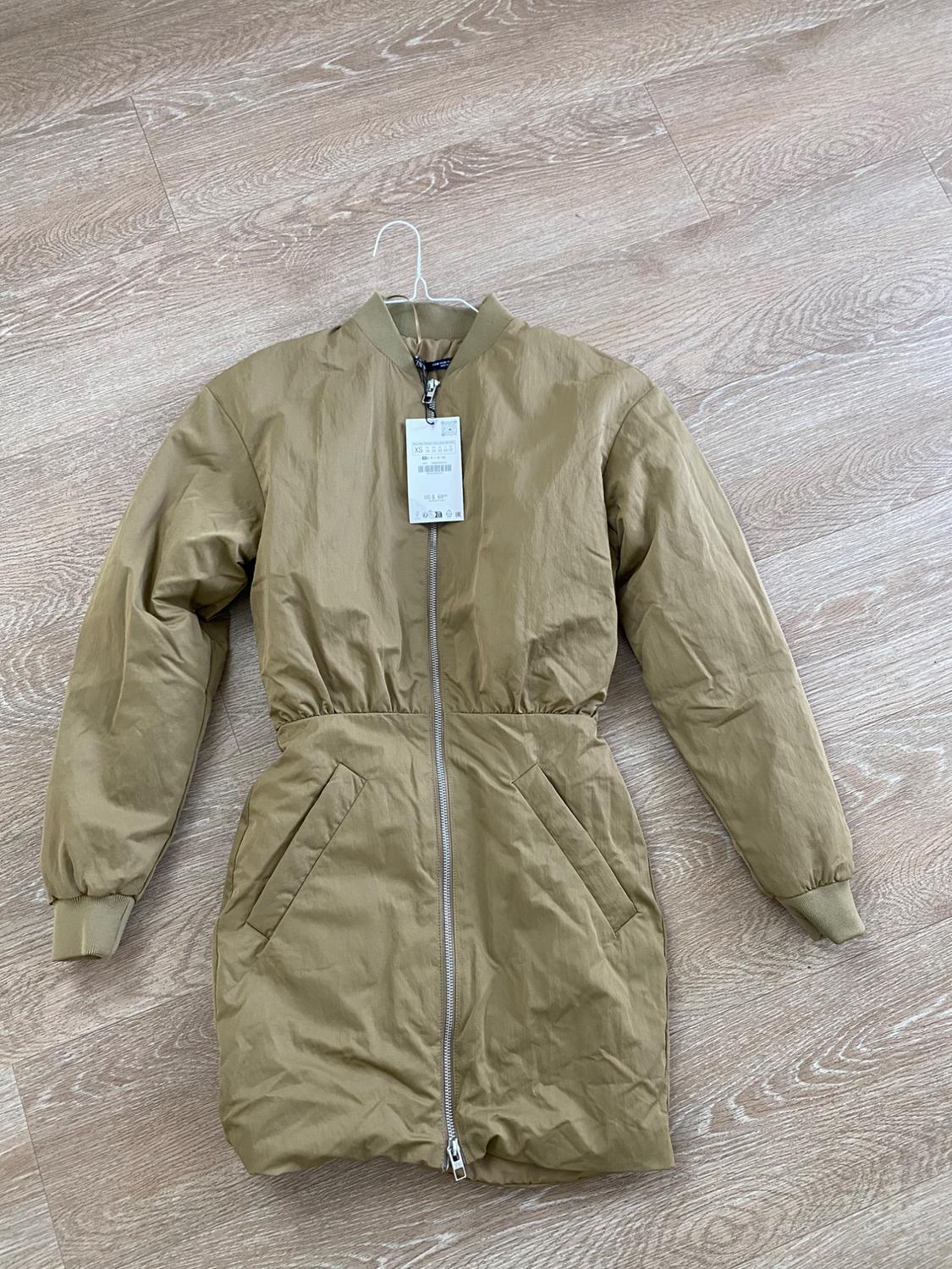 Beige coat with zipper and pockets, hanging on a hanger over light wooden flooring. Visible tag with product details.