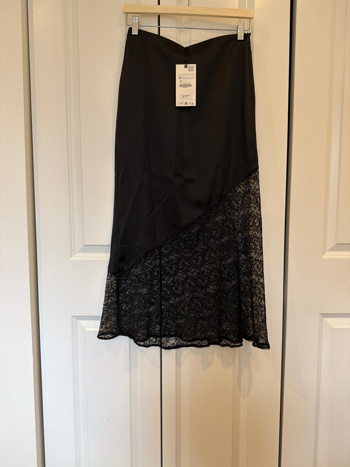 Black skirt hanging on a white door. Smooth top and lace hem with a diagonal cut. Visible label with text XS.