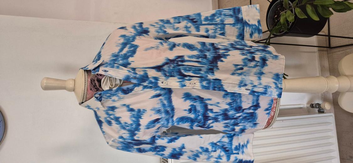 Mannequin in a white long-sleeved shirt with an abstract blue pattern. Indoor background with a potted plant and a white radiator.