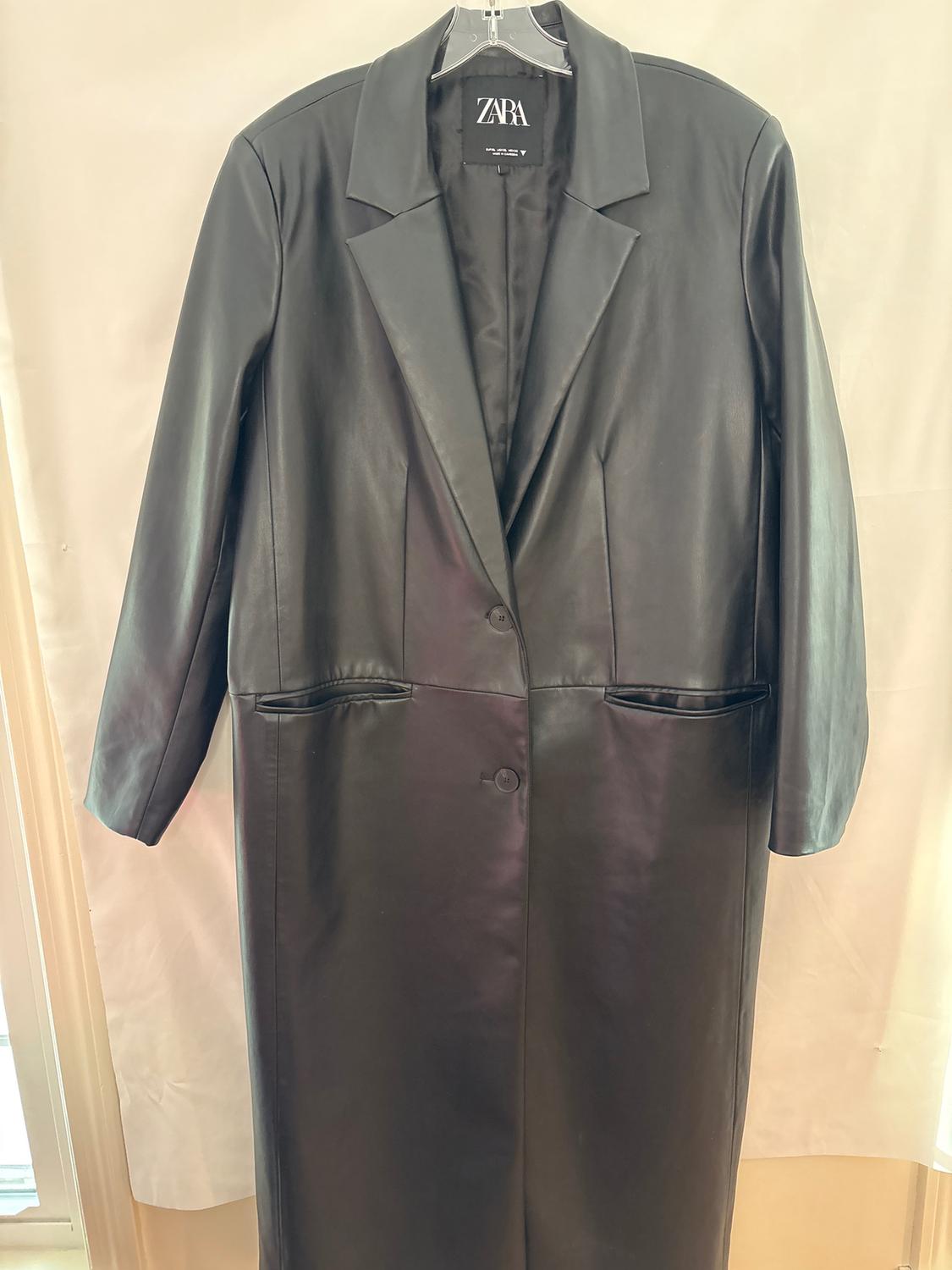 Long black coat hanging on a hanger, with wide lapels and one button. Visible label with the text "ZARA". Light background.