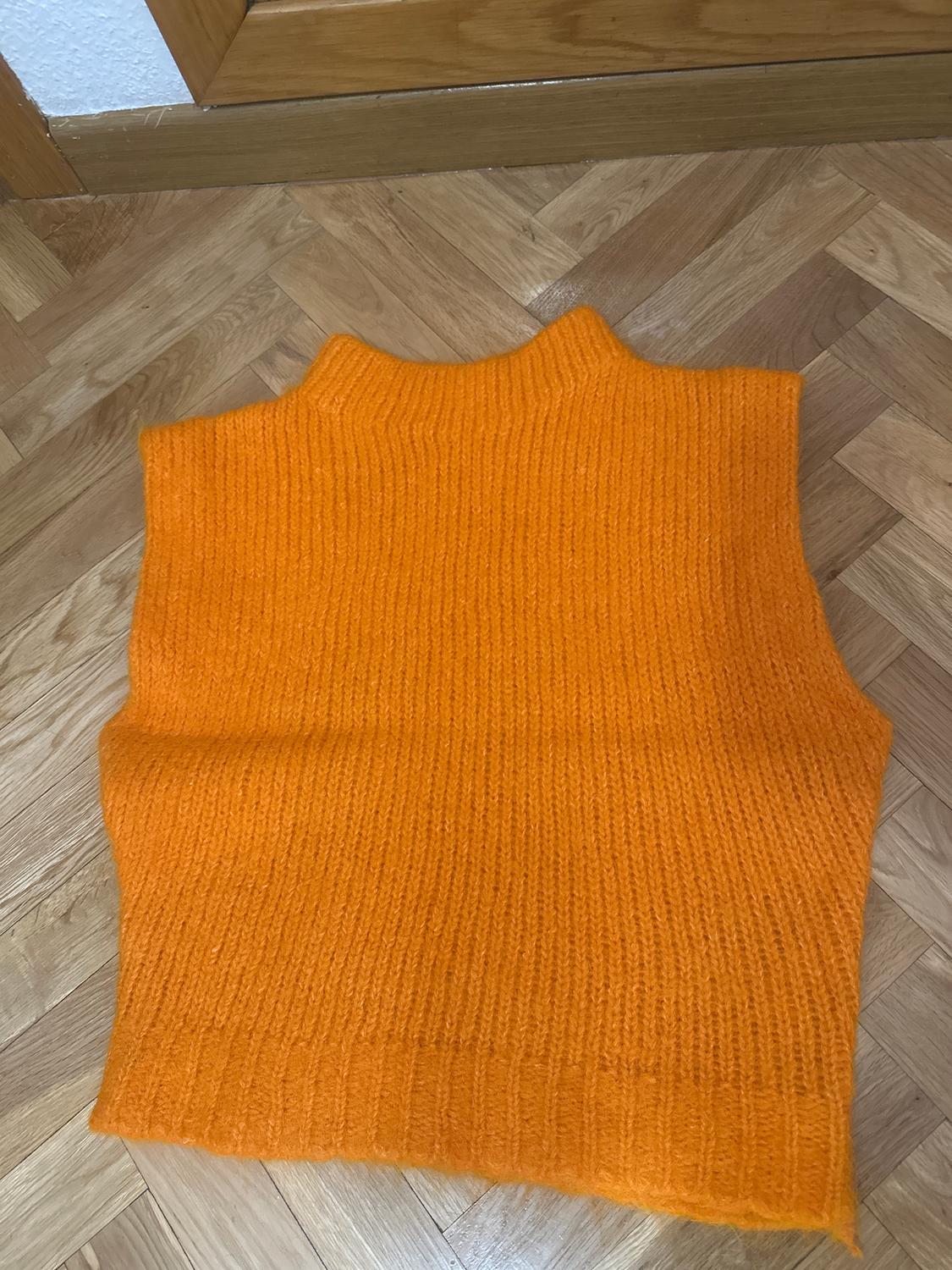 Orange knitted vest on a parquet floor. Sleeveless design with a high neck, highlighting texture and vibrant colour.