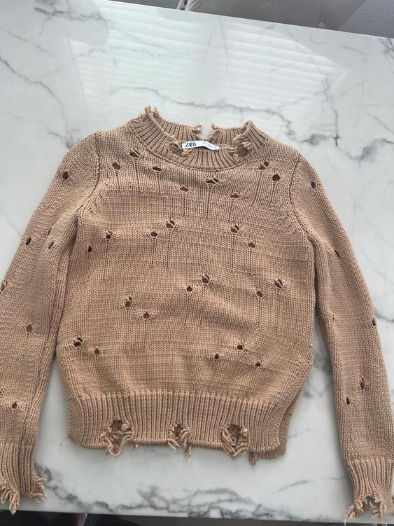 Knit beige sweater with decorative holes and frayed edges on a marble table. Visible label: ZARA.