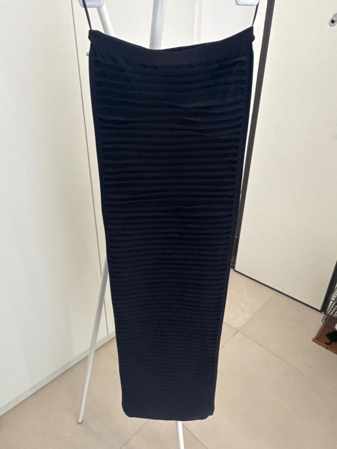 Long black skirt with a horizontal line texture, hanging on a white clothes rail in an interior space with a beige floor and light-coloured walls.