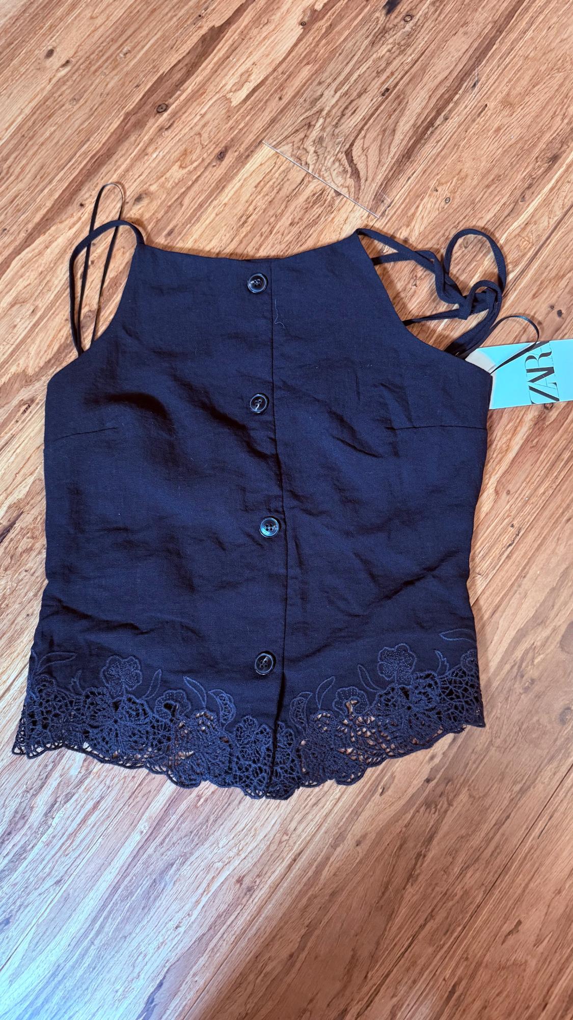 Black spaghetti strap top with front buttons and floral lace hem on a wooden floor. Blue tag with the text ZAR.