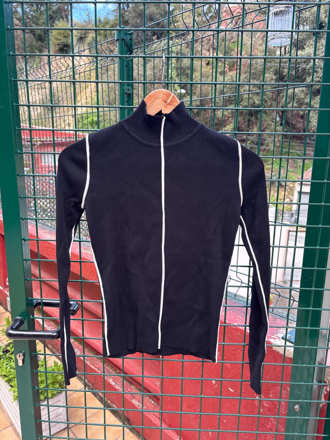 Black high-neck jumper with vertical white stripes, hanging on a wooden hanger over a green fence in an outdoor setting.
