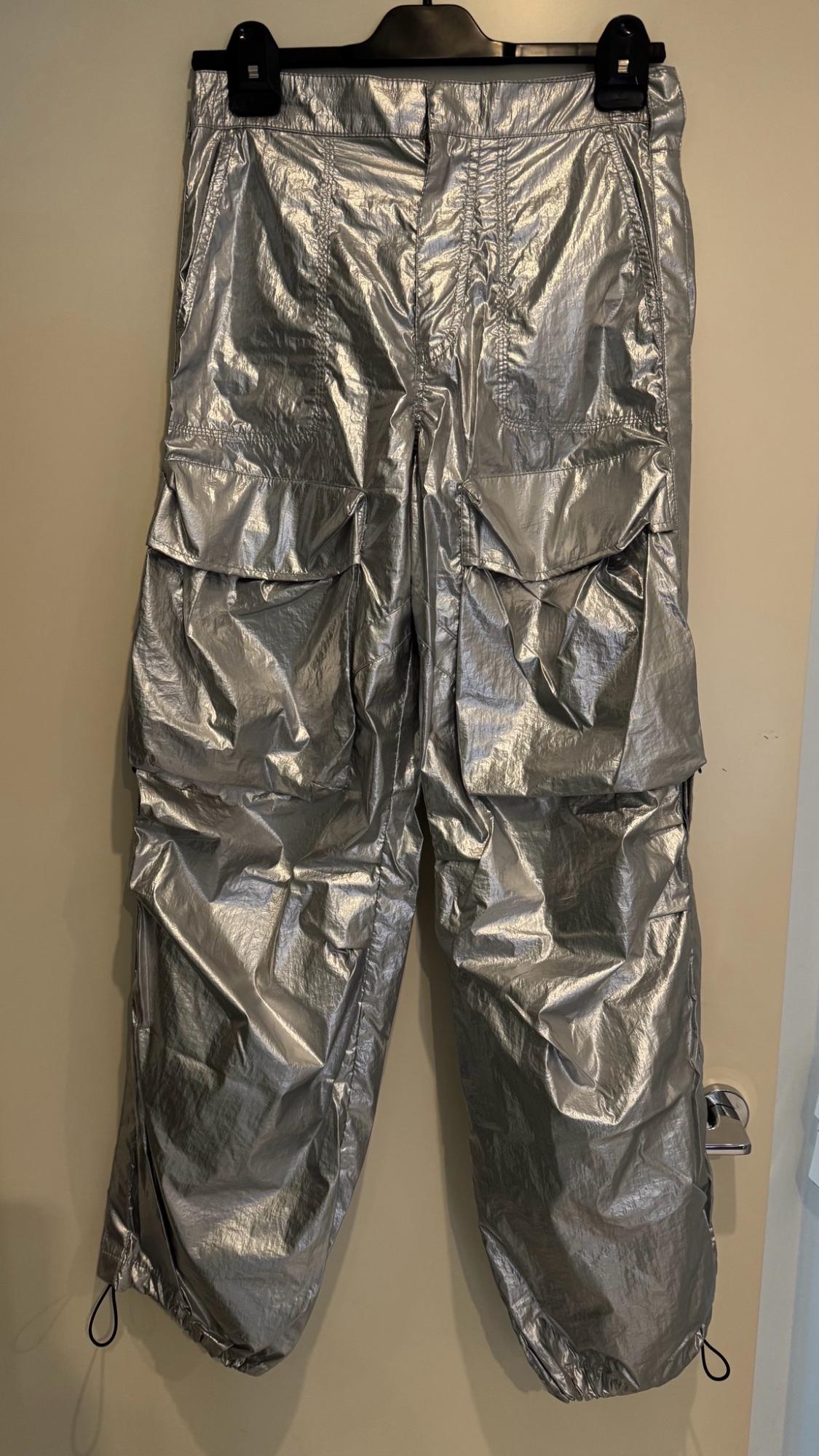 Shiny silver cargo trousers with large pockets and adjustable drawstrings at the hems, hanging from a black hanger on a door.
