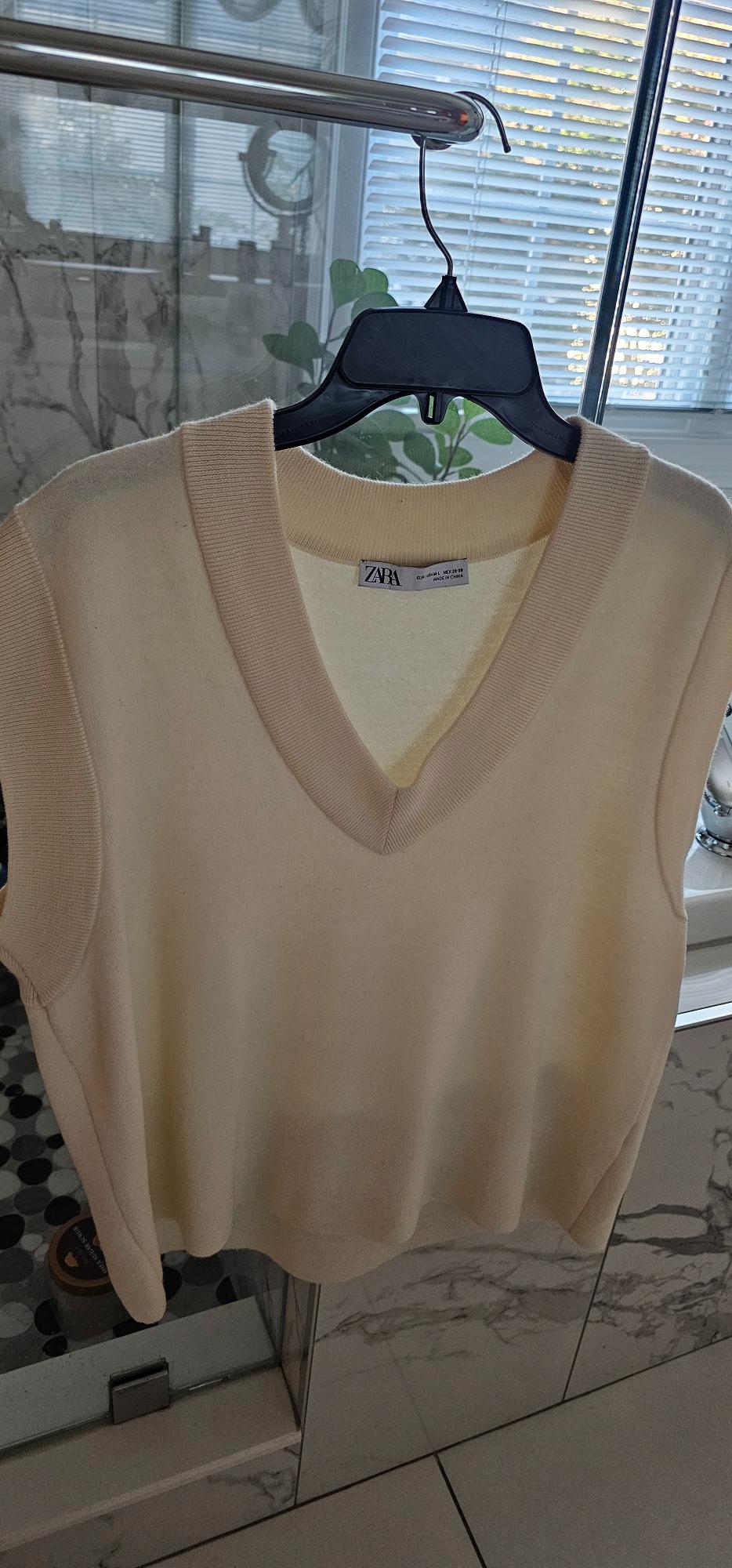 Cream knit vest in a cream color with a V-neck, hanging on a hanger, with a visible Zara brand label inside.
