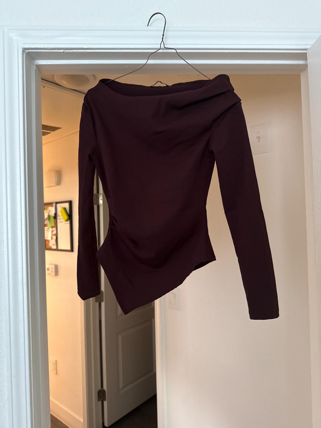 Long-sleeve burgundy top with a draped neckline and an asymmetric hem, hanging on a hanger on a white door frame.