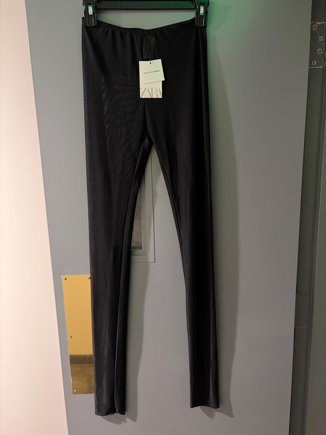 Black pants hanging on a gray door with a label that says "DELICATE GARMENT" and "ZARA". Lit store environment.