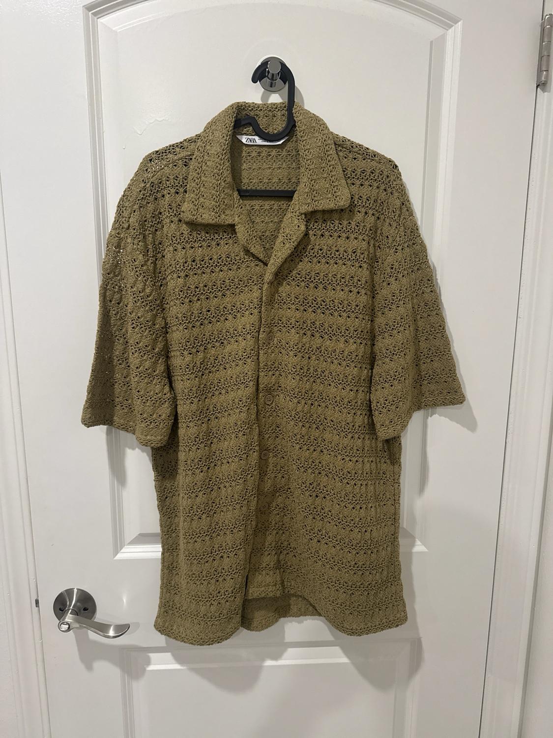 Olive green short-sleeve button-up shirt with an open-knit design, hanging on a white door. Tag with the text ZARA.