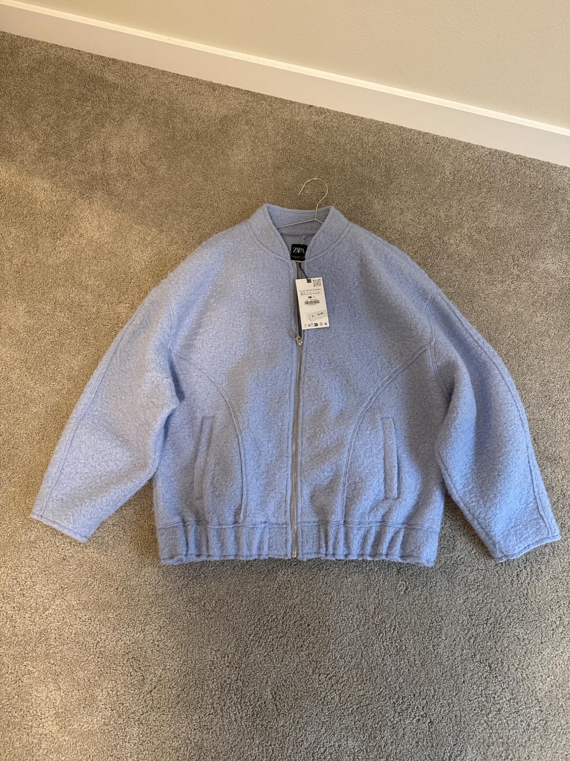 Light blue jacket with zipper and round neck on a gray carpet. Visible label with "ZARA" text and barcode.