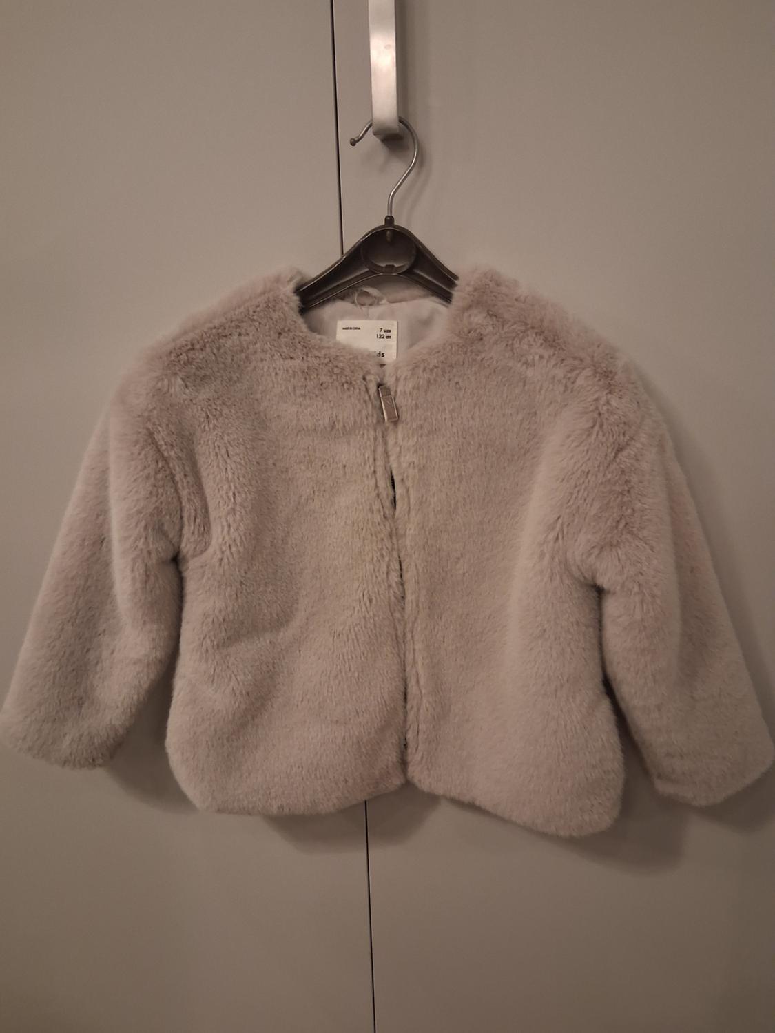 Beige plush jacket hanging on a black hanger over a grey door. Visible label with size 12-18 months.