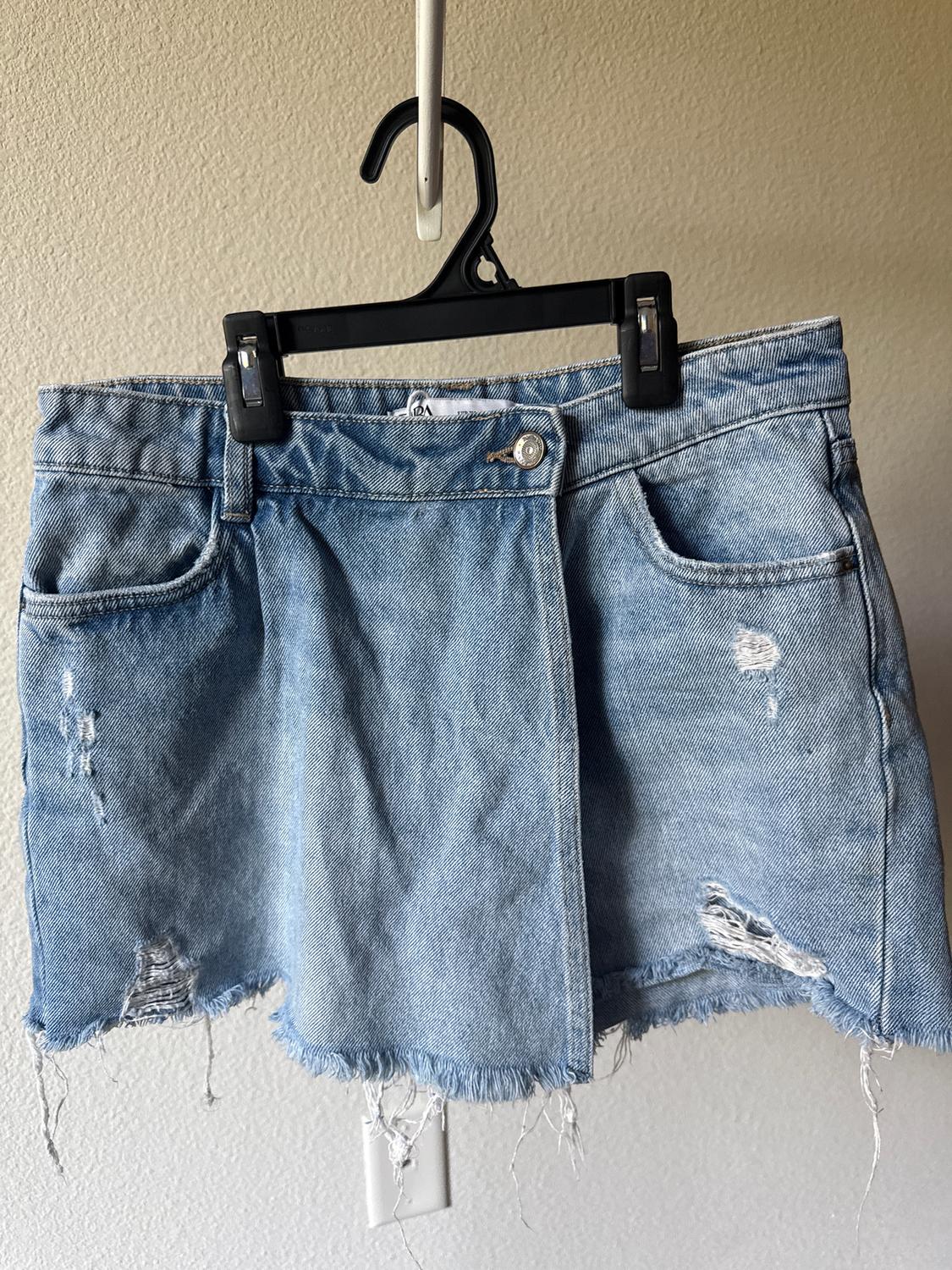 Worn blue denim mini skirt with distressed details and frayed hem, hanging on a black hanger.